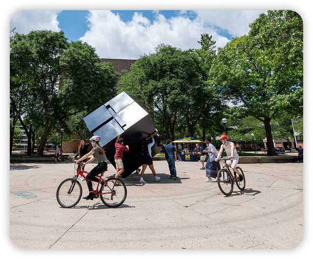 A group of students, some on bicycles, are positioned around the cube on the diag
